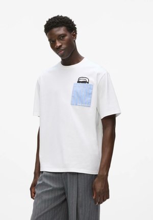 Jeune homme portant un t-shirt blanc oversize avec une poche poitrine rayée bleue et un pantalon large gris à fines rayures, sur fond blanc.