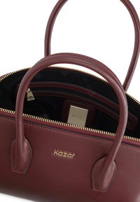 Bordo kožna torba s zlatnim zatvaračem, dvjema ručkama i brendiranim unutarnjim oznakom. Unutrašnjost ima jednu džep sa zatvaračem i obložena je crnim materijalom.