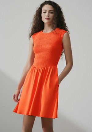 Frau mit langen, lockigen Haaren, die ein ärmelloses, knalloranges Kleid mit einem strukturierten Oberteil und ausgestelltem Rock trägt, steht vor einem schlichten grauen Hintergrund.