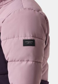 Puffer-Jacke mit einem pinken Oberteil, gerippter Textur und einem lila Unterteil. Ein schwarzes Logopatch befindet sich am Ärmel.