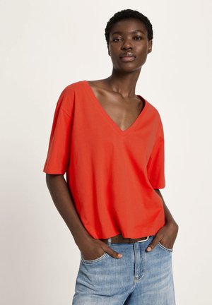 Frau trägt ein locker sitzendes rotes V-Ausschnitt-Kurzarmshirt und hellblaue Jeans, steht mit den Händen in den Taschen vor einem einfarbigen Hintergrund.