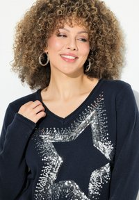 Marineblauer Pullover mit V-Ausschnitt und großem Paillettenstern-Design in Silber. Der Strickstoff weist eine strukturierte Oberfläche und gerippte Bündchen auf.