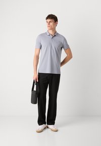 Camisa polo gris claro de mangas cortas, pantalones negros y zapatillas blancas. El modelo sostiene un pequeño bolso negro con una correa.