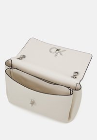 Krémová kožená kabelka přes rameno s otevřeným klopou, která zobrazuje stříbrné "CK" logo a řetězová uchycení, s hlavní komorou a vnitřní kapsou.