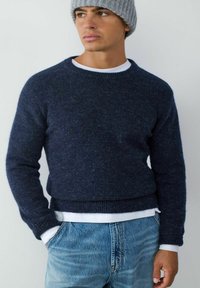 Ciemnoniebieski sweter z dzianiny z prążkowanymi mankietami i dołem, noszony na białej koszuli, zestawiony z jasnoniebieskimi dżinsami.