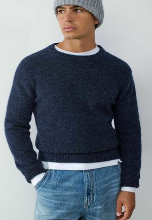 Dunkelblauer Strickpullover mit gerippten Bündchen und Saum, über einem weißen Hemd getragen, kombiniert mit hellblauen Denim-Jeans.