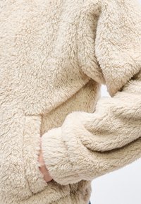 Μπεζ αφράτο φλις μπουφάν με ένα χέρι εν μέρει μέσα σε μπροστινή τσέπη, που δείχνει το μαλακό υφή ύφασμα και τα μακρυά μανίκια με μανσέτα.