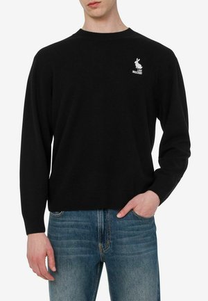 Hombre que lleva un suéter negro de manga larga con un pequeño logo blanco de conejito y el texto "Love Moschino", combinado con vaqueros azules.