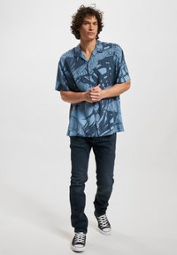 Camicia a maniche corte con bottoni di colore azzurro chiaro, decorata con motivi astratti blu scuro. Abbinata a jeans in denim scuri e scarpe da ginnastica nere.