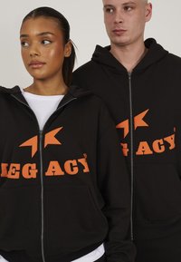 Black zippered hoodies featuring large orange graphics reading "LEGACY" and stars, made of soft fabric with front pockets.