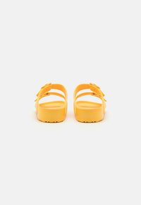 Sandales en plastique jaunes avec un dos ouvert, dotées de trois designs de lanières et d'une semelle texturée pour une meilleure adhérence. Construction simple et légère.