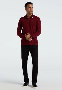 Man in een bordeauxrode polo met lange mouwen, zwarte broek en zwarte sneakers, staand met de handen gekruist, kijkend naar rechts voor een witte achtergrond.