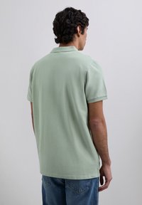 Mies, jolla on tummat kiharat hiukset, pukeutuneena vaaleanvihreään lyhythihaiseen polo-paitaan ja sinisiin farkkuihin, kääntynyt poispäin tasaisen vaaleanharmaan taustan edessä.