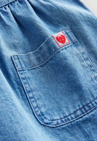 Tissu en denim bleu clair avec un haut froncé, présentant un écusson accentué de rose en forme de fraise sur une poche arrière.