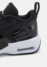 Sneaker noir avec une tige en mesh texturé, des accents en cuir verni brillant, une semelle intermédiaire blanche et une unité de coussin d'air visible.