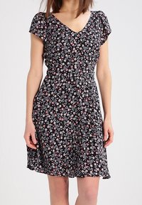 Žena v černých květovaných šatech po kolena s krátkým rukávem a véčkovým výstřihem, stojící na jednom světlém pozadí.