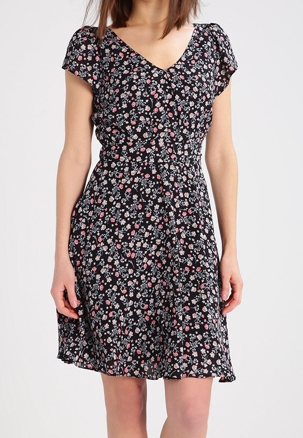 Žena v černých květovaných šatech po kolena s krátkým rukávem a véčkovým výstřihem, stojící na jednom světlém pozadí.