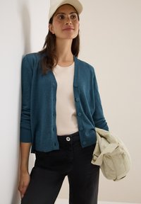 Cardigan teal avec un col rond, une fermeture à boutons et des poignets côtelés. Associé à un haut clair et un pantalon noir, tenant un sac de couleur claire.