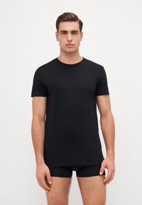 T-shirt noir à manches courtes en coton, avec un col rond et une coupe près du corps. Le modèle porte des sous-vêtements noirs.