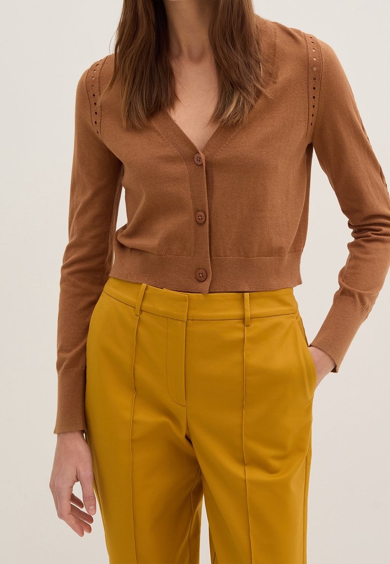 Cardigan corto marrón con escote en V, mangas texturizadas con detalles de ojal y tres botones marrones en el frente, combinado con pantalones mostaza.
