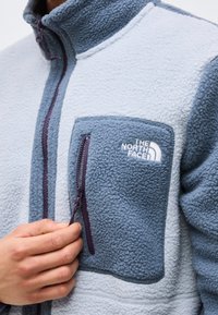 Hand, die den Reißverschluss einer blau-grauen Fleecejacke mit The North Face-Logo auf der Brusttasche hält.