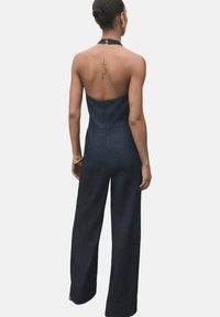 Jeans jumpsuit med figurnära liv och vida byxor, med halterneck och rygglös design. Mörk tvätt och jämn textur.