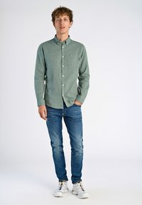 Camisa verde de manga larga con botones y bolsillo en el pecho, combinada con jeans ajustados azules y zapatillas deportivas blancas con detalles coloridos.