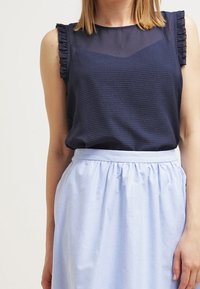 Mörkblå ärmlös topp med texturerad mönster och volangdetaljer på axlarna, ihop med en ljusblå, plisserad knälång kjol.