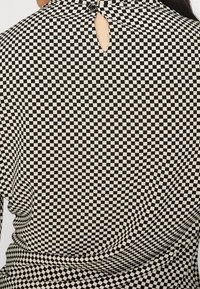 Černobílý kostkovaný top s knoflíkovým zapínáním na zádech. Tkanina má hladkou strukturu a je doplněna o detail výřezového klíče.