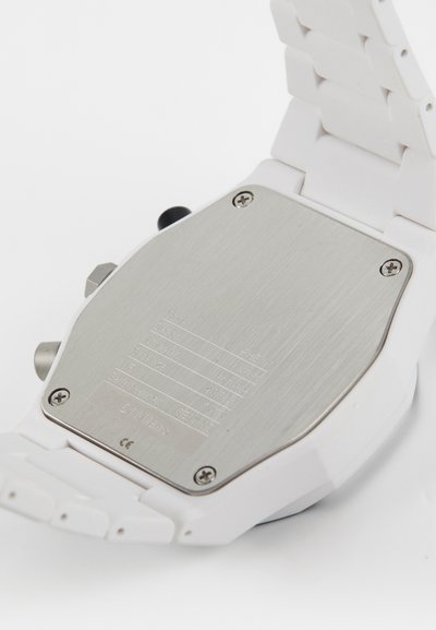 Hvid armbåndsur med en glat plastikrem og bagside i rustfrit stål. Har skruer og indgraverede specifikationer på bagsiden.