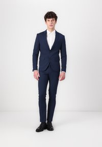 Marineblauer Anzug aus strukturiertem Stoff mit einer taillierten Passform. Besitzt zwei Knöpfe, einen Reverskragen und seitliche Taschen, kombiniert mit einem weißen Hemd.