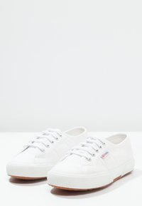 Sneaker bianche in tela con suole in gomma, caratterizzate da un profilo basso, punta rotonda e cinque occhielli metallici per i lacci. Leggera texture e design semplice.