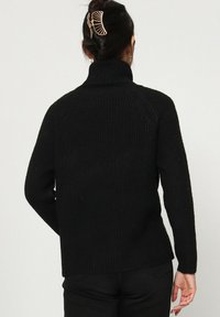 Femme aux cheveux bruns en chignon, portant un pull noir à col roulé et un pantalon noir, tournant le dos à un fond clair.