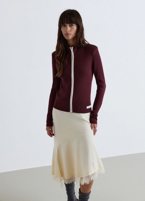 Woman wearing a ribbed maroon zip-up sweater and cream midi skirt with lace hem standing against a plain white background.