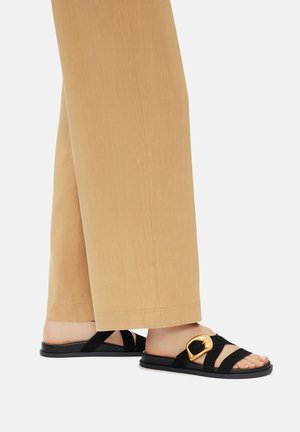 Czarne klapki na płaskiej podeszwie z szeroką złotą klamrą. Posiadają krzyżujące się paski i płaską czarną podeszwę, zestawione z jasnobrązowymi spodniami o szerokich nogawkach.