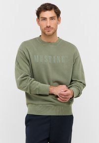 Sweatshirt vert olive avec texture côtelée et col rond, arborant un logo "MUSTANG" en relief de couleur assortie sur la poitrine.