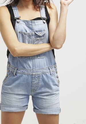 Salopette en denim bleu clair avec shorts roulés, présentant des bretelles ajustables, une poche avant et des accents de boutons sur les côtés.