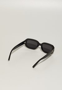 Black rectangular sunglasses with dark lenses, thick plastic frame, subtly curved temples, and gold hardware detail on the side.
