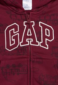 Bordó babakapucnis pulóver fekete vonatillusztrációkkal, nagy fehér kontúrral körülvett "GAP" felirattal elöl és középen cipzárral.