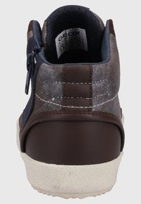 Zapatilla de cuero y textil marrón con un collar acolchado, cremallera trasera y acentos en azul marino contrastantes. Suela de goma texturizada con borde blanco.