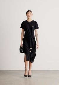 Μαύρο φόρεμα με ζώνη κόμπο στη μέση και ανοίγματα στο πλάι, κατασκευασμένο από απαλό ύφασμα. Συνδυασμένο με μαύρες γόβες και μια μικρή, υφασμάτινη τσάντα.