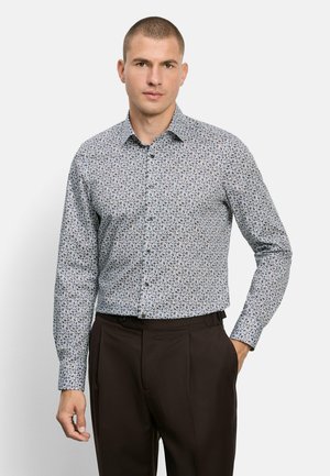 Mann trägt ein langärmliges, geknöpftes Hemd mit Blumenmuster und dunkle Hose, steht mit einer Hand in der Tasche vor einfachem Hintergrund.