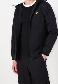 Zwarte lichtgewicht jas met capuchon, ritssluiting, elastische manchetten en een klein geel logo op de borst, gedragen over een zwart T-shirt met ronde hals.