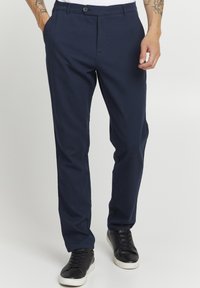 Marine skreddersydde bukser laget av glatt stoff, med en frontknee og to sidelommer, kombinert med sorte sneakers.
