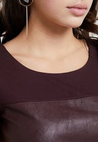 Haut bordeaux en tissu lisse avec un panneau en cuir. Le design comprend un col rond et des détails de couture subtils.