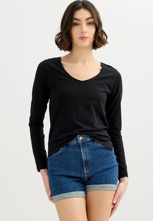 Joven mujer con camiseta negra de manga larga con escote en V y pantalones cortos de mezclilla azul remangados, de pie frente a un fondo claro y sencillo.