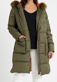 Olivengrüner, wattierter Parka mit Kapuze aus Kunstfell, Frontreißverschluss, zwei großen Aufsetztaschen und kontrastierendem orangefarbenem Innenfutter.