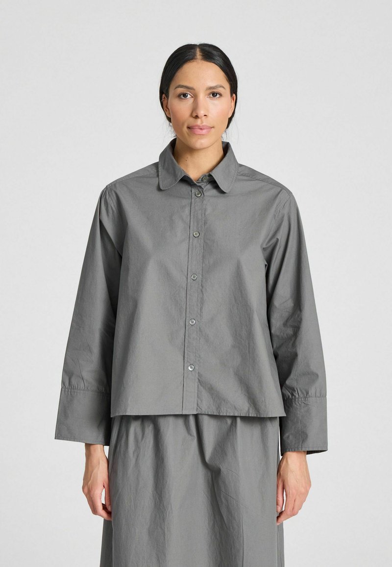 Camicia grigia a maniche lunghe, con colletto piatto e taglio dritto. Il tessuto sembra morbido, con una leggera lucentezza e minima texture.