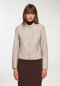 Giacca di pelle beige con un design strutturato, dotata di una chiusura con zip dorata, colletto alto e maniche lunghe, abbinata a un vestito marrone.