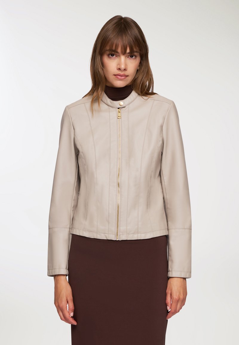 Giacca di pelle beige con un design strutturato, dotata di una chiusura con zip dorata, colletto alto e maniche lunghe, abbinata a un vestito marrone.
